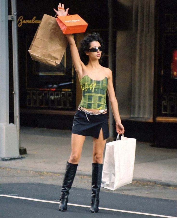 a woman in black boots and a green shirt carrying shopping bags on the street with her hand up