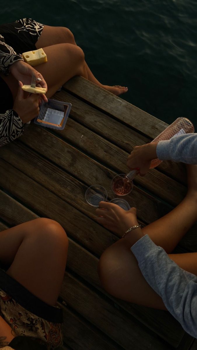 three people sitting on a dock holding wine glasses and cell phones in their hands as they eat