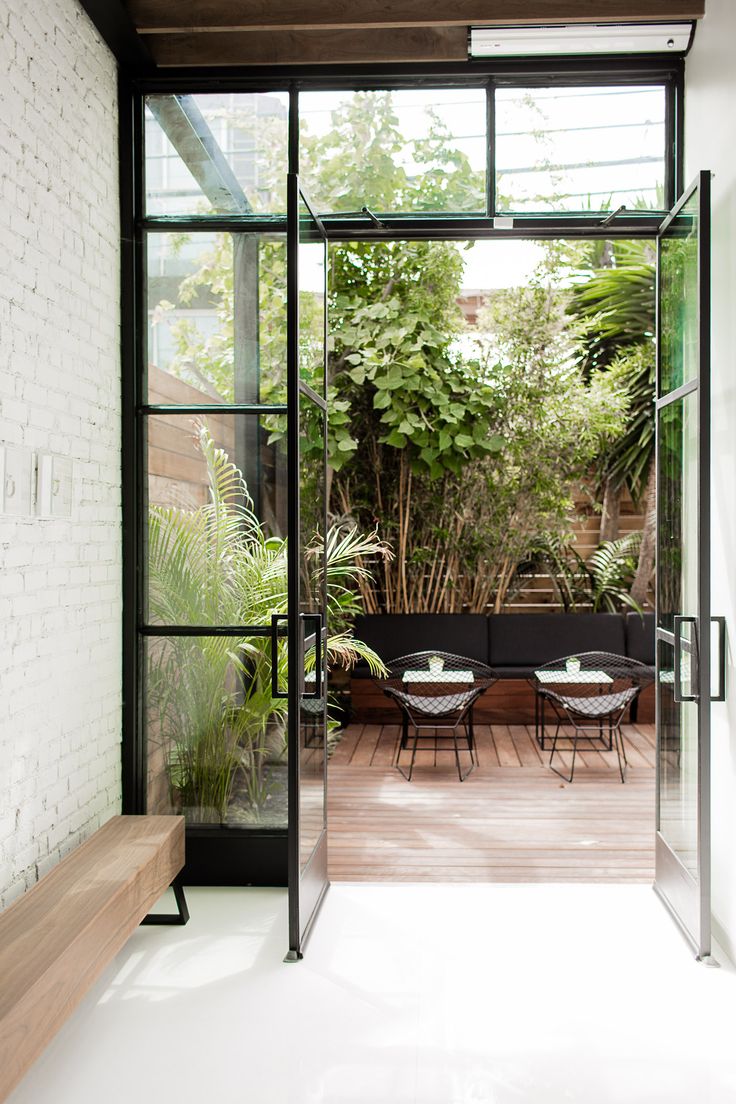 an open glass door leading to a patio area with tables and chairs on it, surrounded by greenery