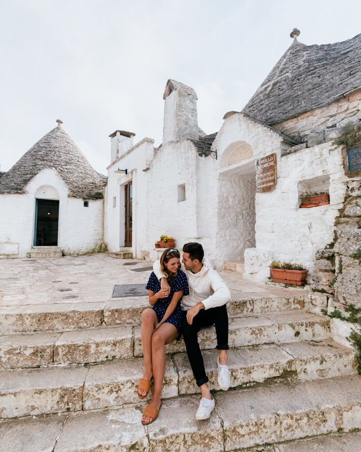 a man and woman sitting on steps in front of an old building with thatched roof