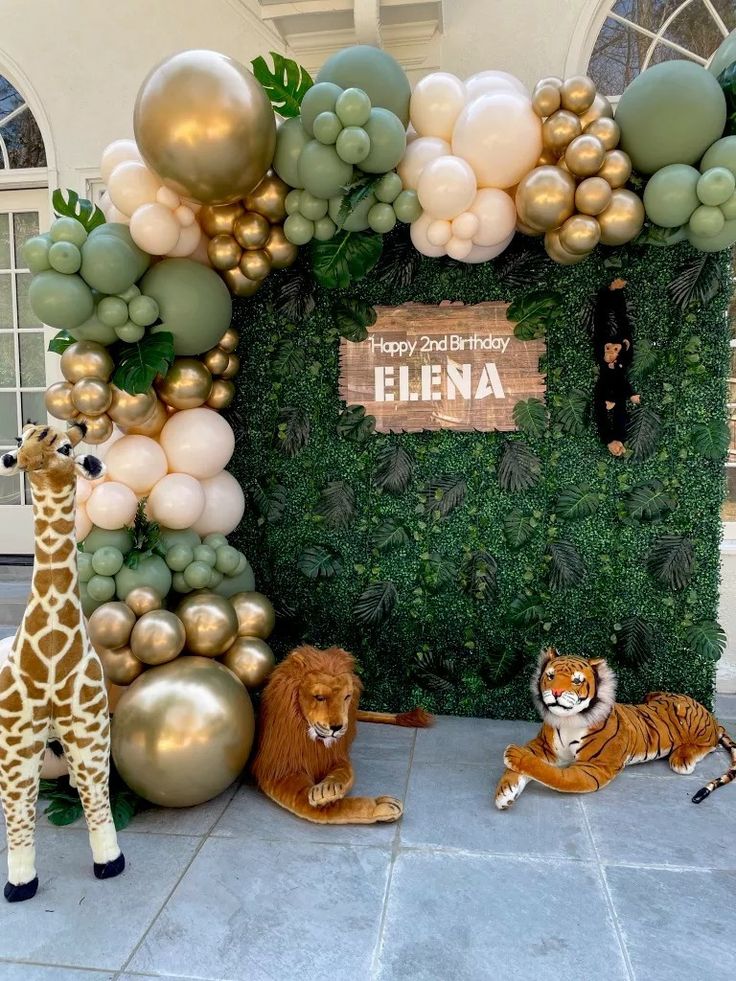 two stuffed animals are sitting in front of a backdrop with balloons and greenery for an animal themed birthday party