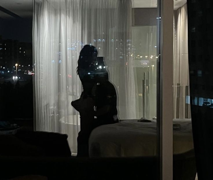 a person standing in front of a window with curtains on the outside and city lights behind them