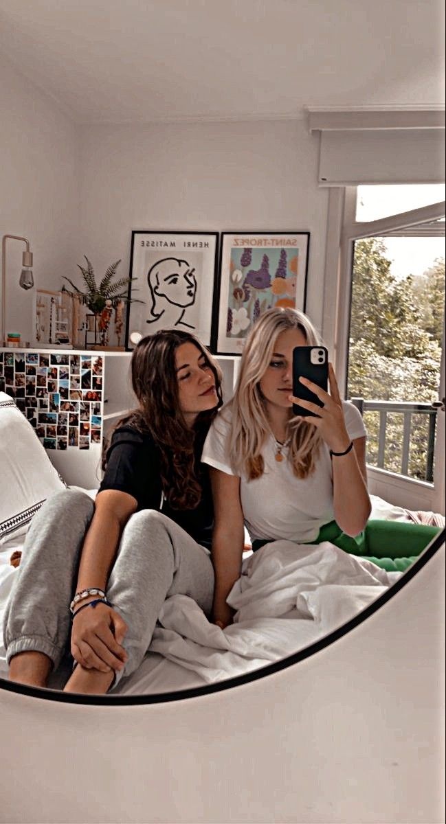 two women sitting on a bed looking at their cell phones and taking pictures in the mirror