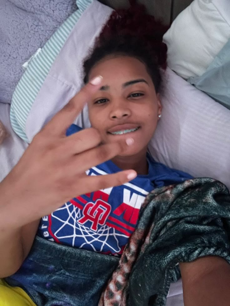 a woman laying on top of a bed holding her hand up to the camera and making a peace sign