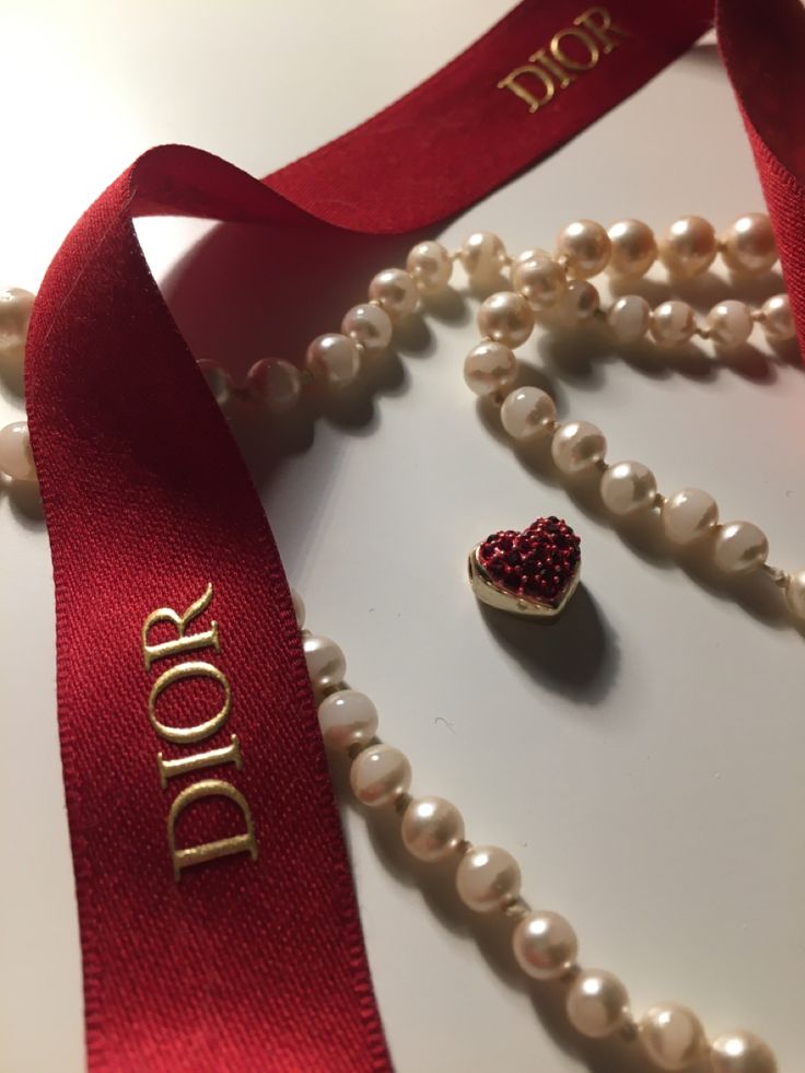 a necklace with pearls and a heart shaped brooch sits on a white surface next to a red ribbon