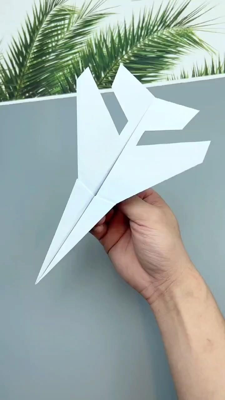 a hand holding up a paper airplane in front of a plant on a gray wall