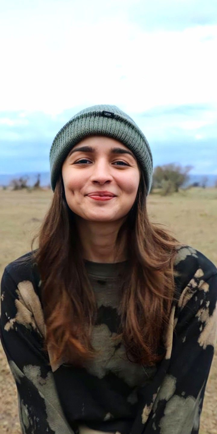 a woman with long hair wearing a green beanie smiles at the camera while sitting in a field