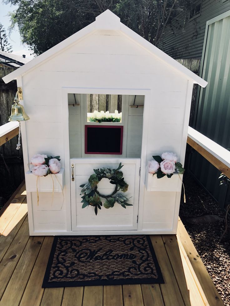 a small white house with wreaths and flowers on the front door is sitting on a wooden deck