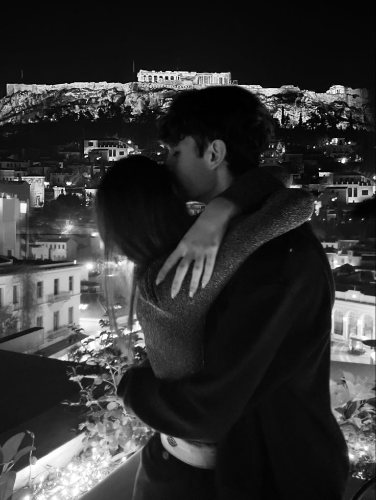 a man and woman kissing in front of a cityscape at night with lights on