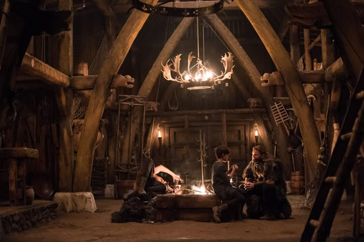 three people sitting around a fire pit in a wooden building with lights hanging from the ceiling