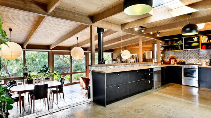 an open kitchen and dining room area in a house with wood ceilinging, large windows, black cabinetry, and red rug on the floor