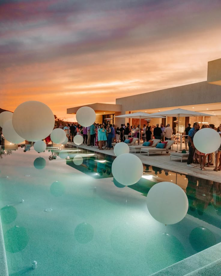 people are gathered around an outdoor pool at sunset or dawn with white balloons floating in the air