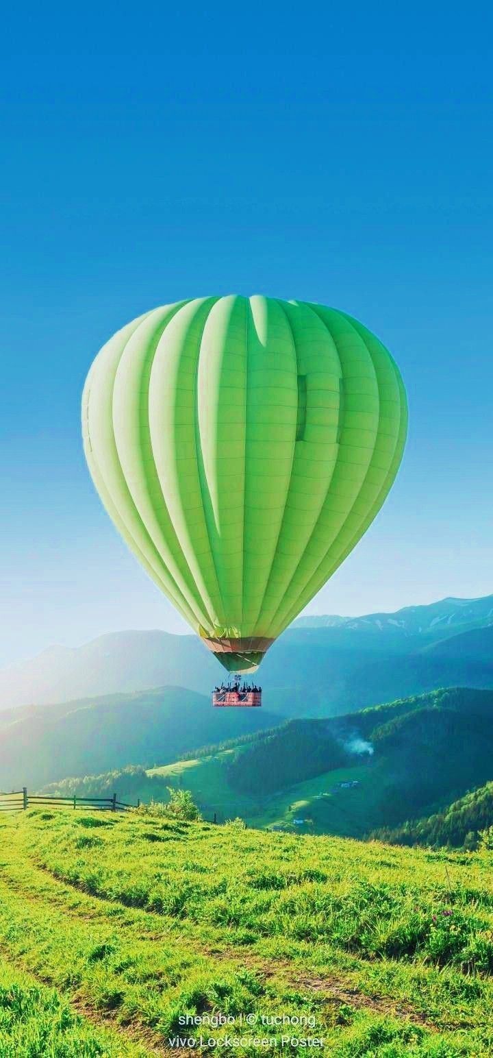 a green hot air balloon flying over a lush green hillside under a blue cloudless sky