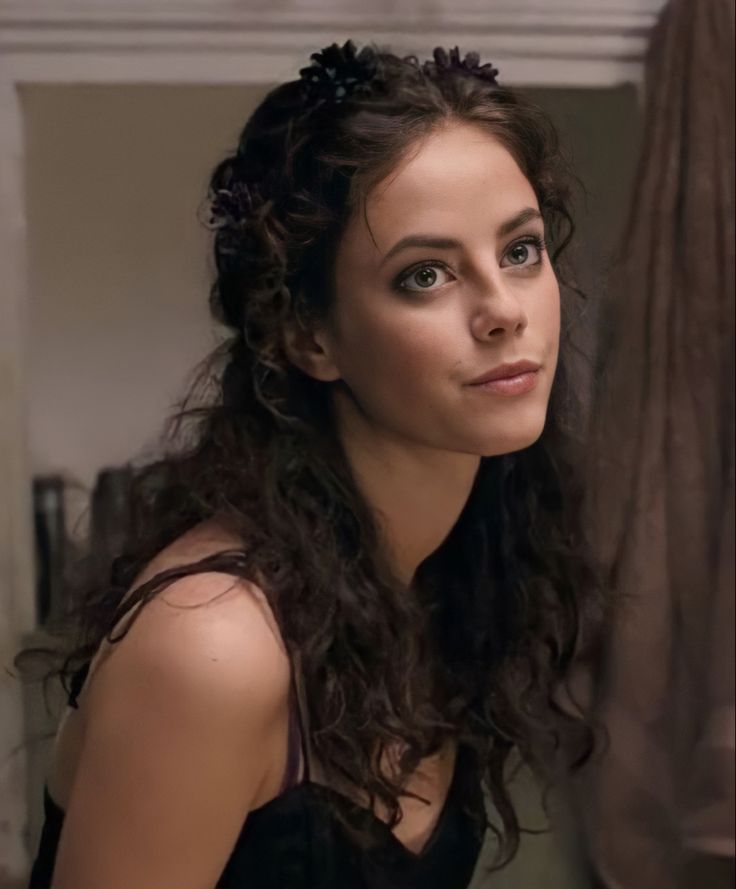 a woman with long curly hair looking into the distance while wearing a black dress and flower in her hair