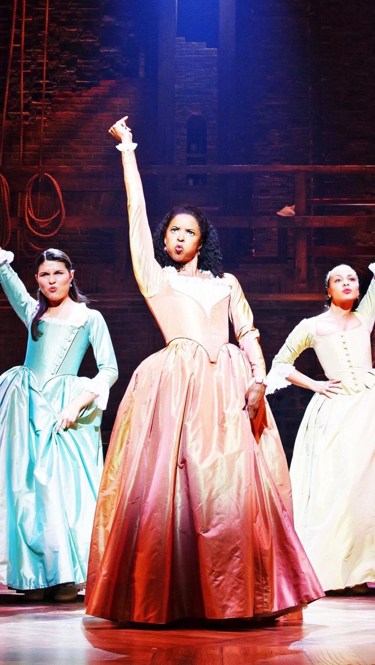 three women in long dresses on stage with their arms up and one woman holding her hand out