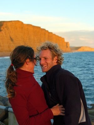 a man and woman standing next to each other near the ocean with cliffs in the background