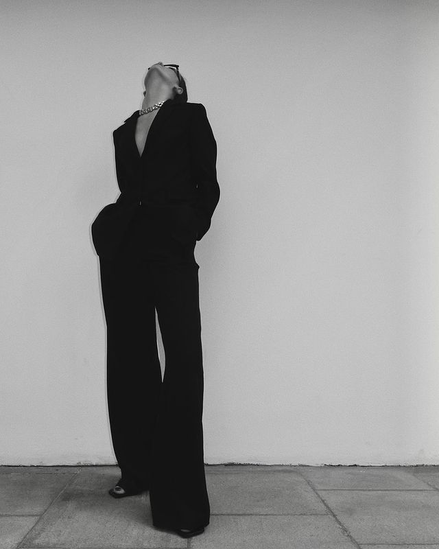 a woman in a black suit leaning against a wall with her hands on her head