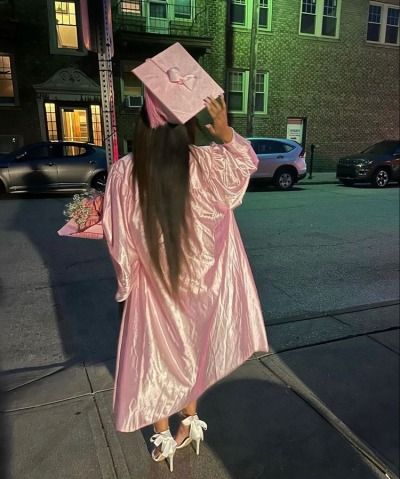 a woman in a pink graduation gown is walking down the street with her long hair pulled back