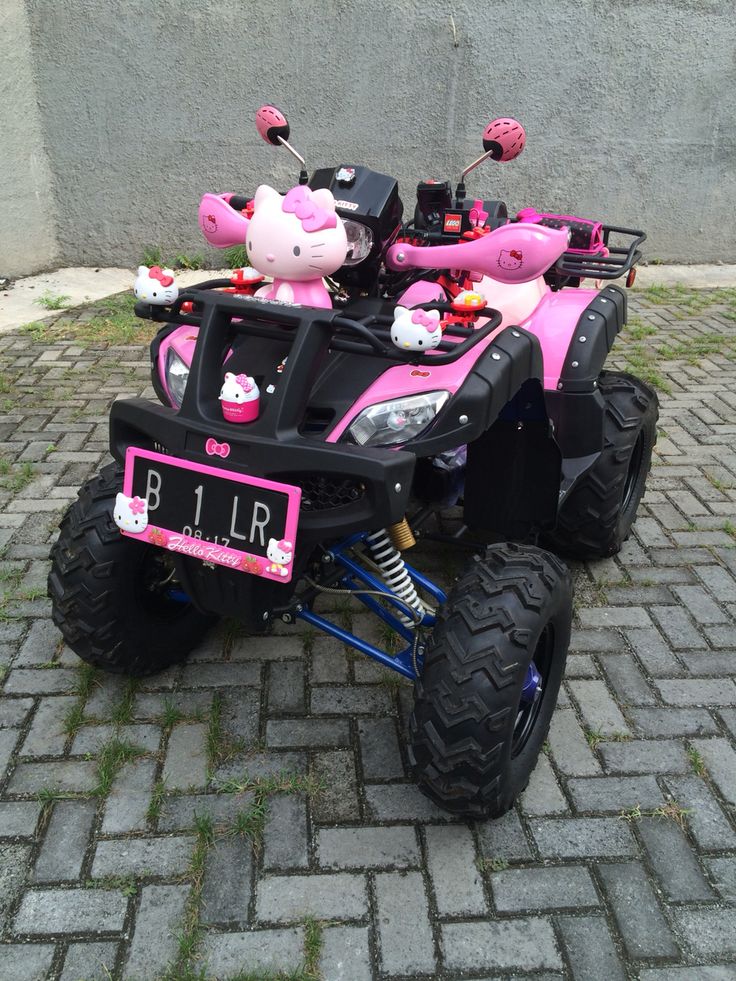 a pink toy atv with hello kitty decorations on the front and back wheels, parked on a brick sidewalk