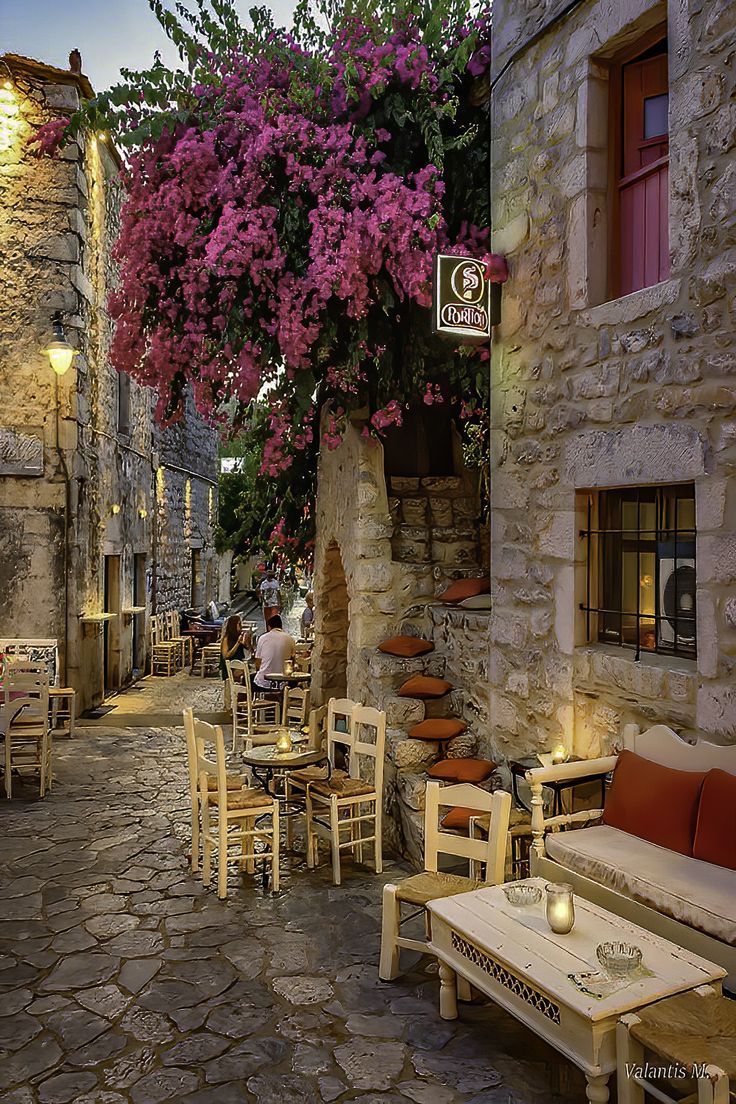 an alleyway with tables and chairs lined up against the stone wall, surrounded by purple bougaia