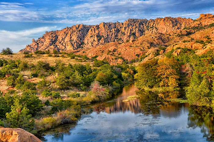 a river running through a lush green forest next to a rocky mountain range with clouds in the sky