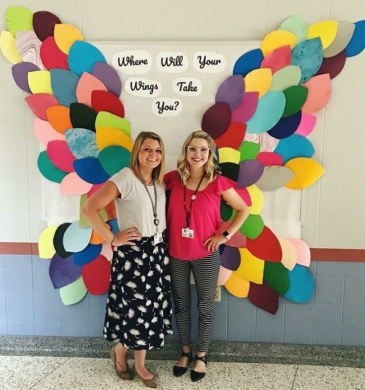 two women standing next to each other in front of a wall with colorful paper butterflies