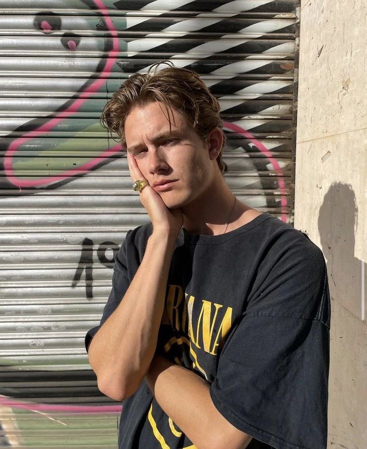 a young man leaning against a wall with his hand on his chin and looking at the camera