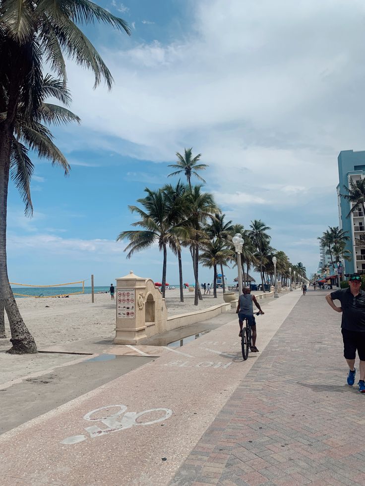 two people riding bikes on the beach near palm trees and buildings with blue sky in background