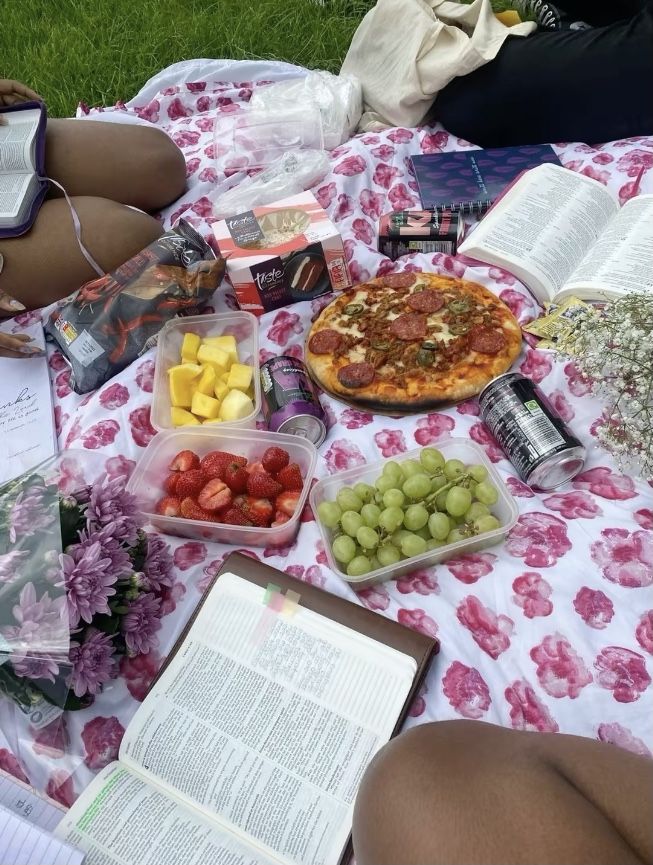 people are sitting on the grass with food and books in front of them, while one person is reading a book