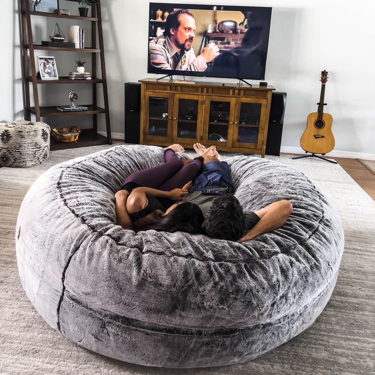 two people laying on a giant bean bag chair in front of a flat screen tv