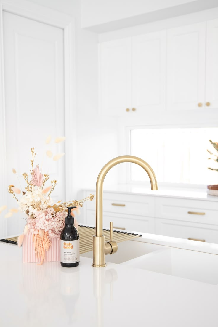 a white kitchen with gold faucet and flowers on the counter top, next to a bottle of shampoo