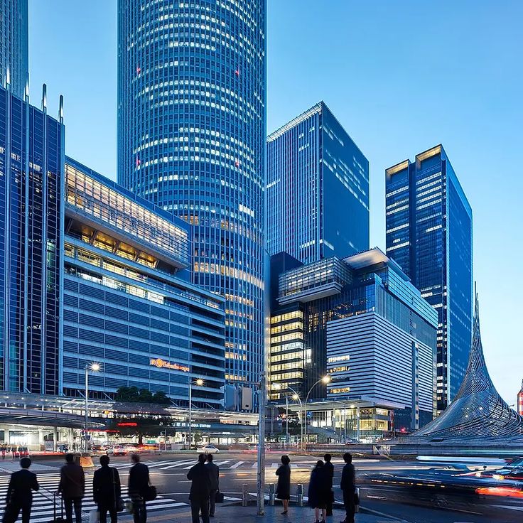 many people are standing on the sidewalk in front of some tall buildings at night time