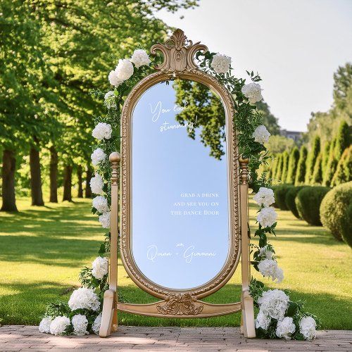 a large mirror sitting on top of a sidewalk next to white flowers and greenery