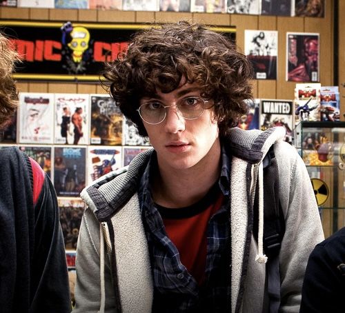 a young man with curly hair and glasses looks at the camera while standing in front of comic books