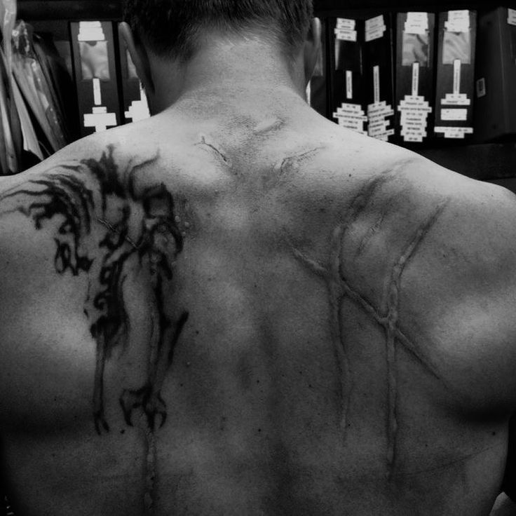 the back of a man with tattoos on his upper and lower half, standing in front of bookshelves