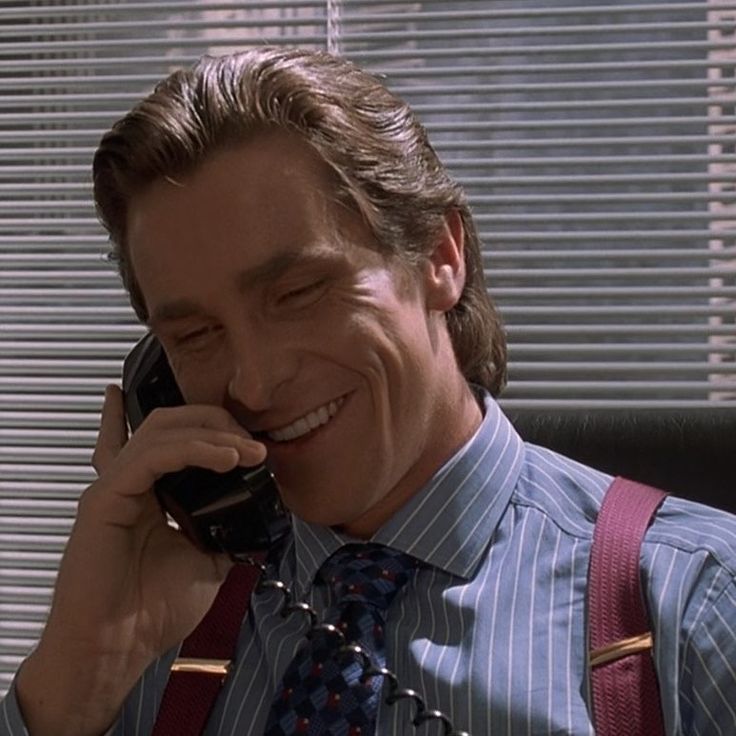 a man in suspenders talking on a cell phone while sitting at a desk with his eyes closed