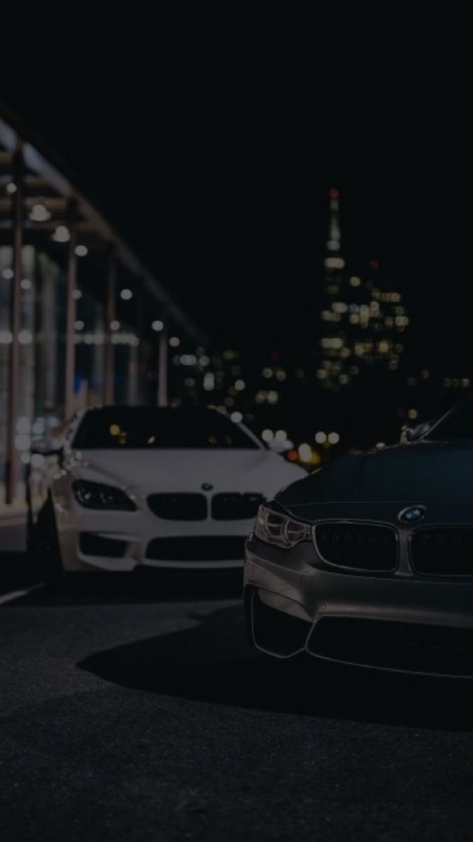 two cars parked in front of a tall building at night with the city lights behind them