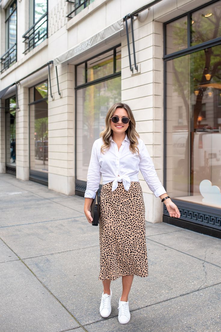 Fashion and lifestyle influencer Jessica Rose Sturdy wearing a cropped white shirt with a leopard skirt and white tennis shoes for work. White Sneakers Work Outfit Summer, Styling Dress With Sneakers, Summer White Shirt Outfit, White Sneakers Skirt Outfit, White Trainers With Dress, Dress With Sneakers Work Outfit, White Shoes And Dress Outfit, Crop Shirt With Skirt, White Printed Skirt Outfit