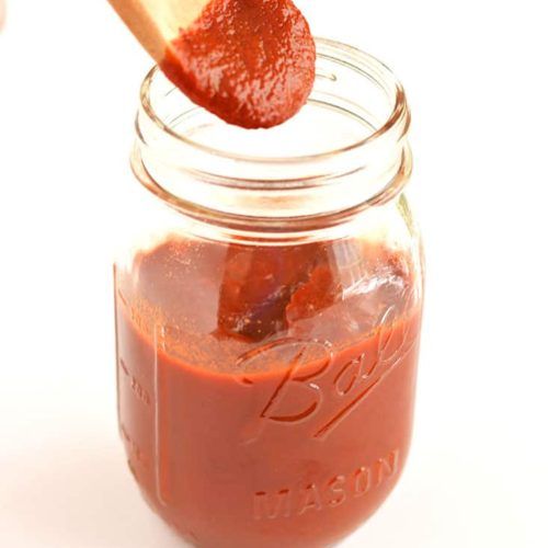 a spoon full of tomato sauce being held up by a jar with it's lid