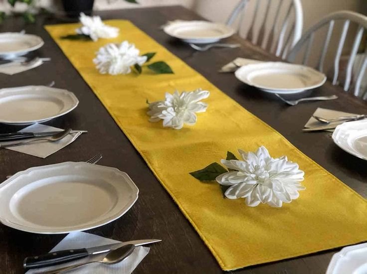 the table is set with plates, silverware and white flowers on yellow cloth runner