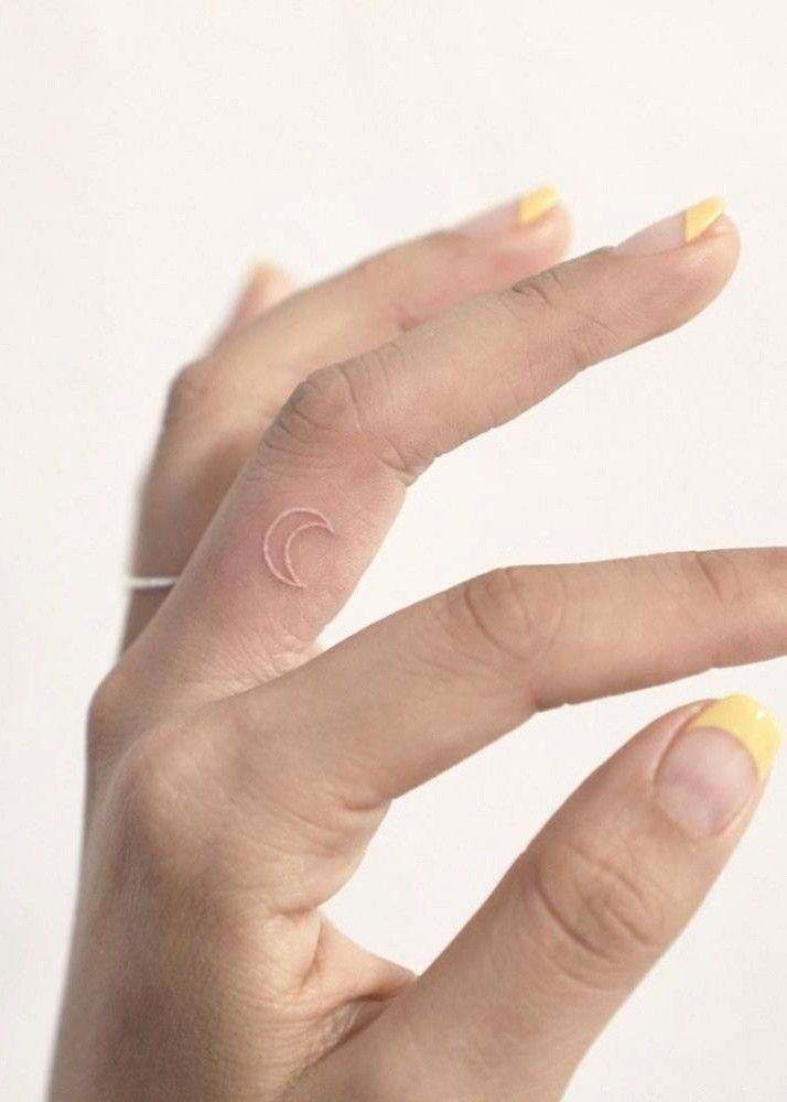 a person's left hand with yellow nail polish and a small crescent tattoo on the middle finger