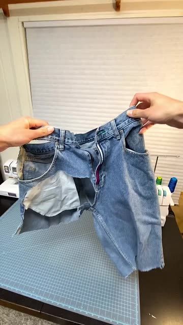 two hands hold up a pair of jeans that have been folded on top of a table