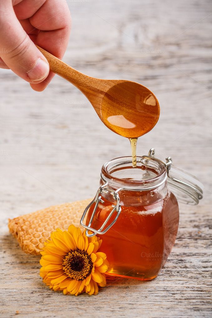 a wooden spoon pouring honey into a jar with a yellow flower on the table next to it