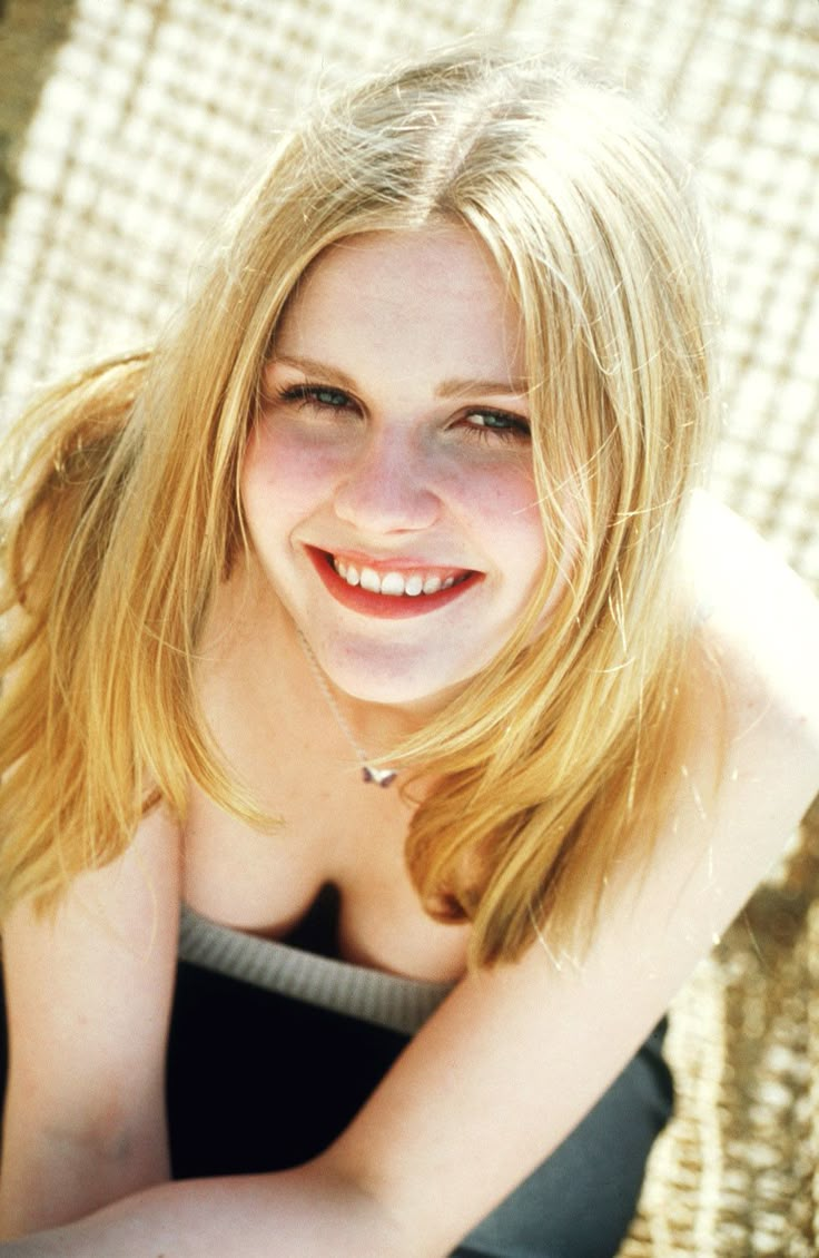 a woman with blonde hair sitting on a chair smiling and looking at the camera while wearing a black tank top