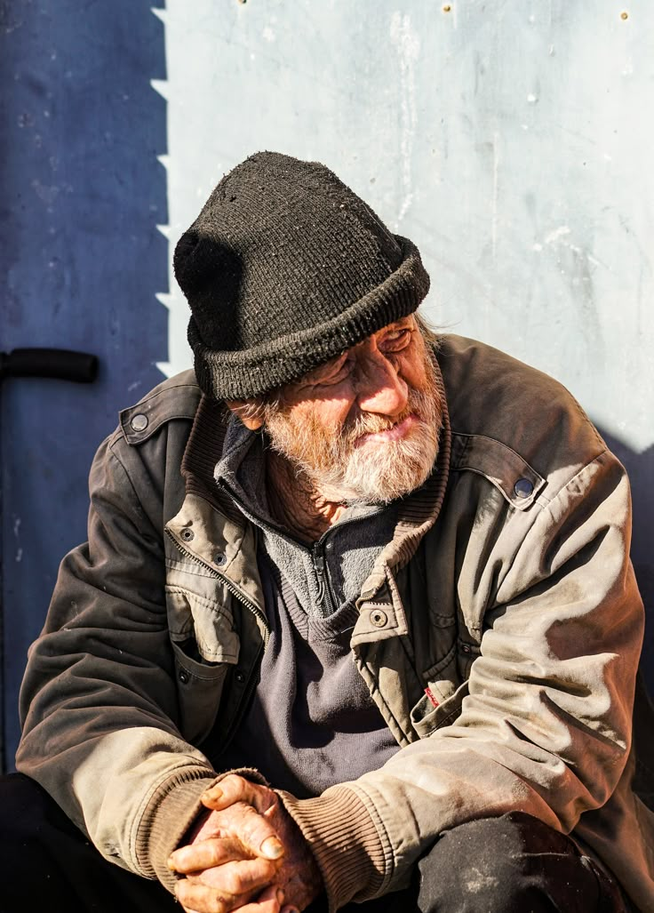 an old man wearing a black hat sitting on the ground with his hands clasped to his knees