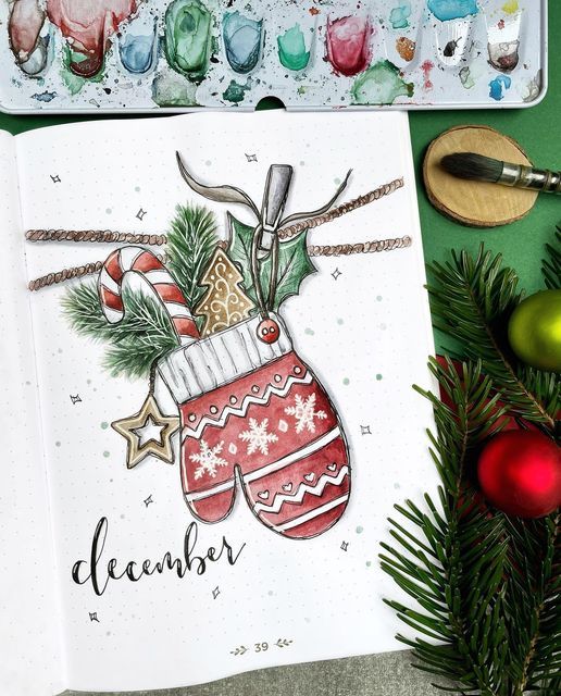 an open notebook with christmas stockings on it next to some ornaments and paintbrushes