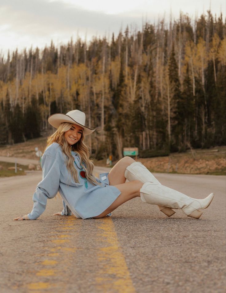 a woman wearing a cowboy hat and dress sitting on the side of a road with trees in the background