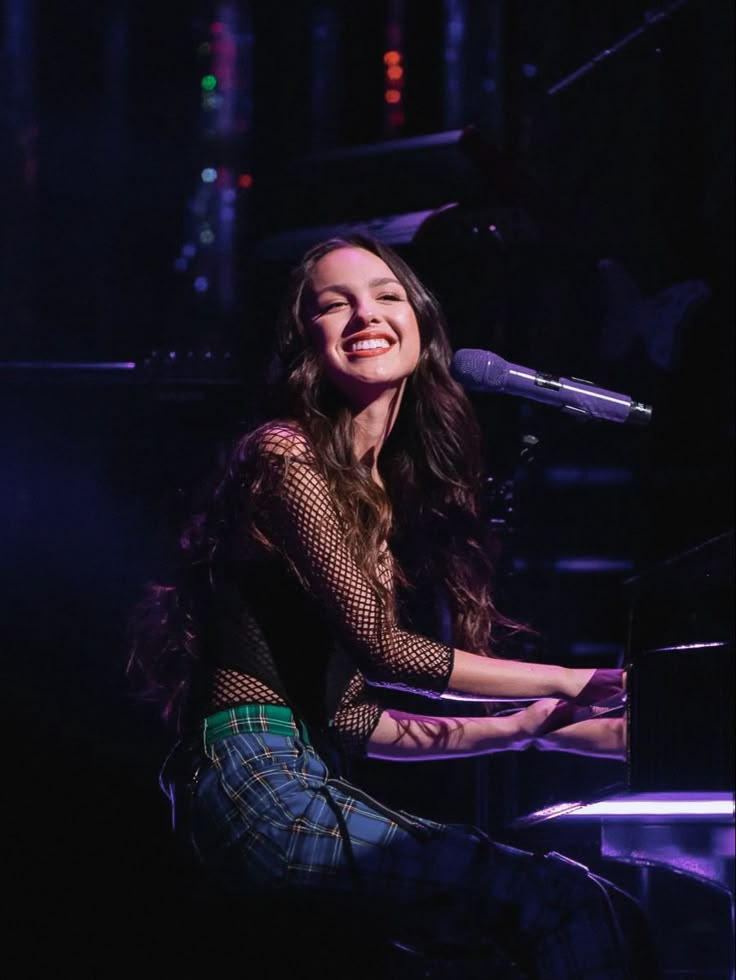 a smiling woman sitting at a piano in front of a microphone and purple light behind her