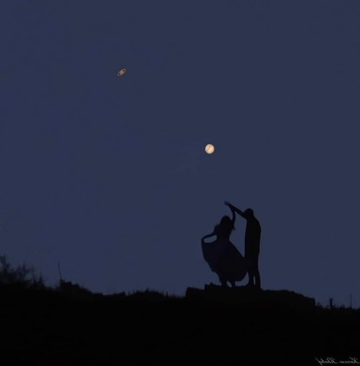 two people standing on top of a hill at night with the moon in the background