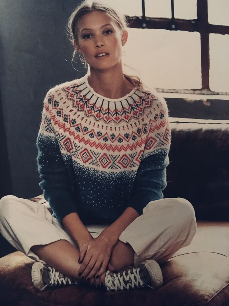 a woman sitting on top of a couch in front of a window wearing a sweater